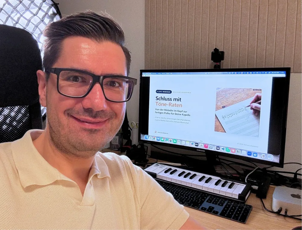 Daniel Käsbauer lächelnd am Arbeitsplatz, hinter ihm auf dem Bildschirm die Startfolie des Live-Webinars